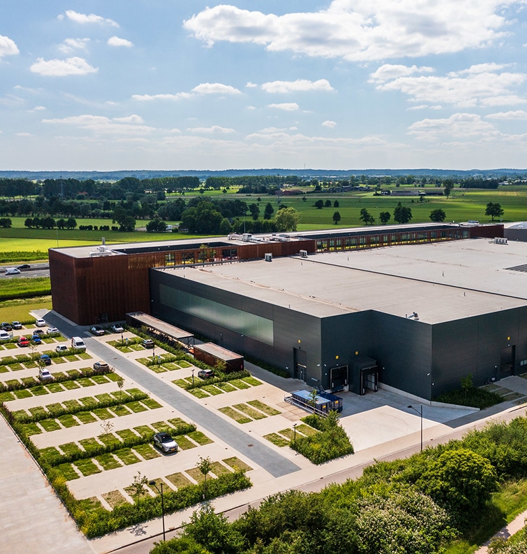Aerial view of a large commercial building, in Amsterdam, with a flat roof, surrounded by a parking lot with several cars, green fields, trees, and a partly cloudy sky in a rural setting