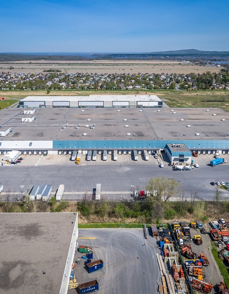 Aerial view of a large industrial warehouse complex with multiple loading docks, parked trucks and trailers, equipment and containers in the foreground, and a residential area with houses and fields in the background under a clear blue sky