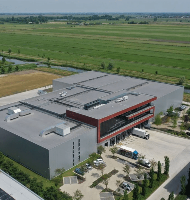 Aerial view of a modern industrial building in Amsterdam, with a flat roof and red section featuring large windows, surrounded by green fields, trees, vehicles, paved roads, and agricultural land in the distance