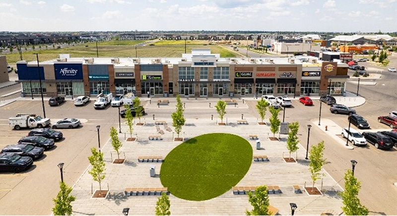 a bird eye view of strip filled with stores and a vehicle filled parking lot. Nestled in the cent is a brick lined seating area with benched and a green pace.