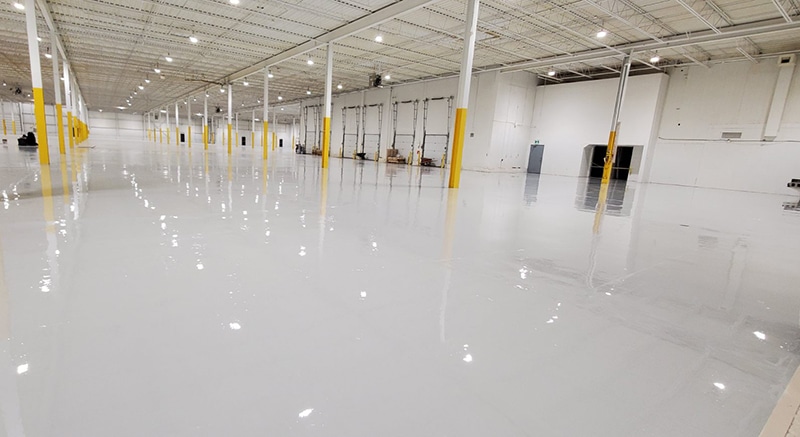 Large empty warehouse with a polished reflective floor, white columns with yellow bases, high ceiling with evenly spaced lights, and pallets and equipment near the wall