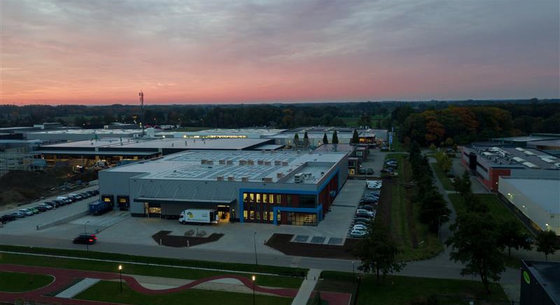 Aerial view of a modern industrial complex at dusk, featuring a flat-roofed building with skylights and ventilation units, adjacent parking lot with cars, surrounding green spaces, and roadways under a partly cloudy sky with pink and orange sunset hue