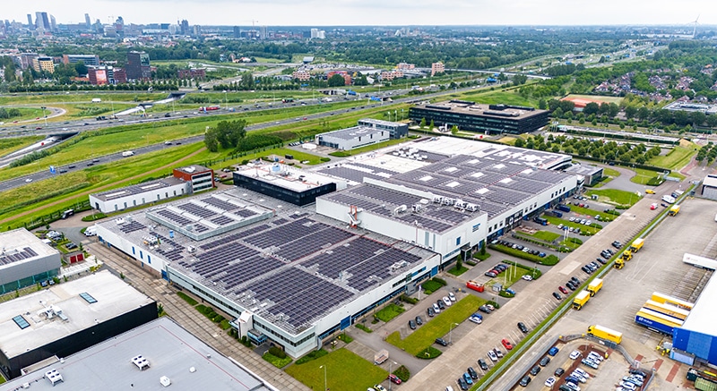 Aerial view of an industrial area with large buildings featuring rooftop solar panels, surrounded by parking lots, green spaces, roads, and a highway with vehicles in the background