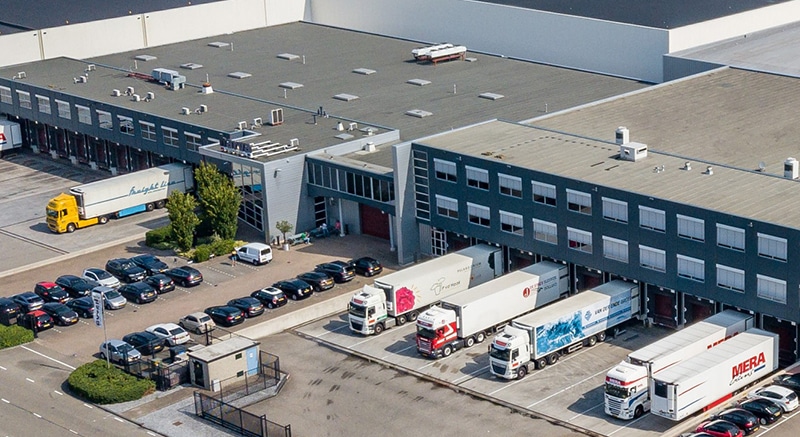 Aerial view of a large industrial warehouse with multiple loading docks, trucks being loaded or unloaded, a flat roof with equipment, and adjacent parking lot filled with cars