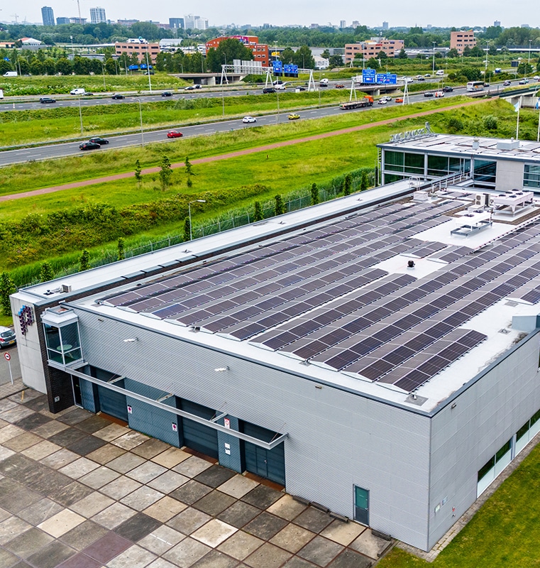 Large industrial building with a flat roof covered in solar panels, located near a multi-lane highway and surrounded by green spaces, office buildings, and residential areas under an overcast sky